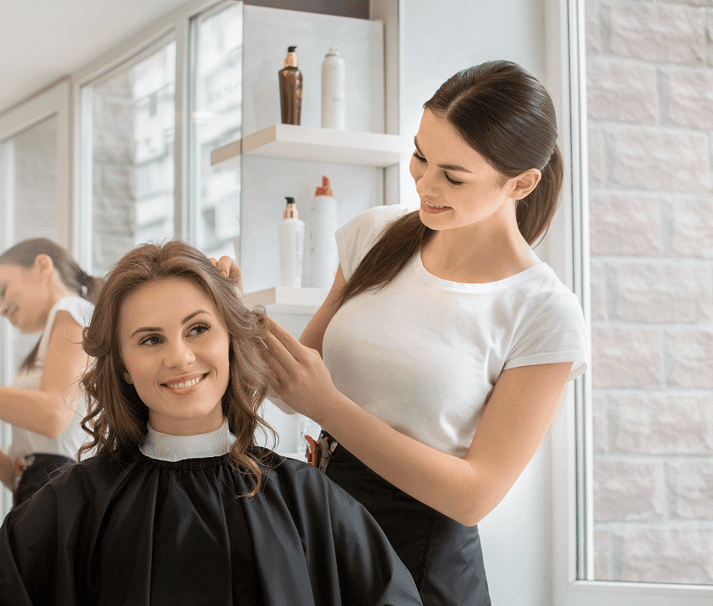 A stylist combing a happy woman's hair at a salon.